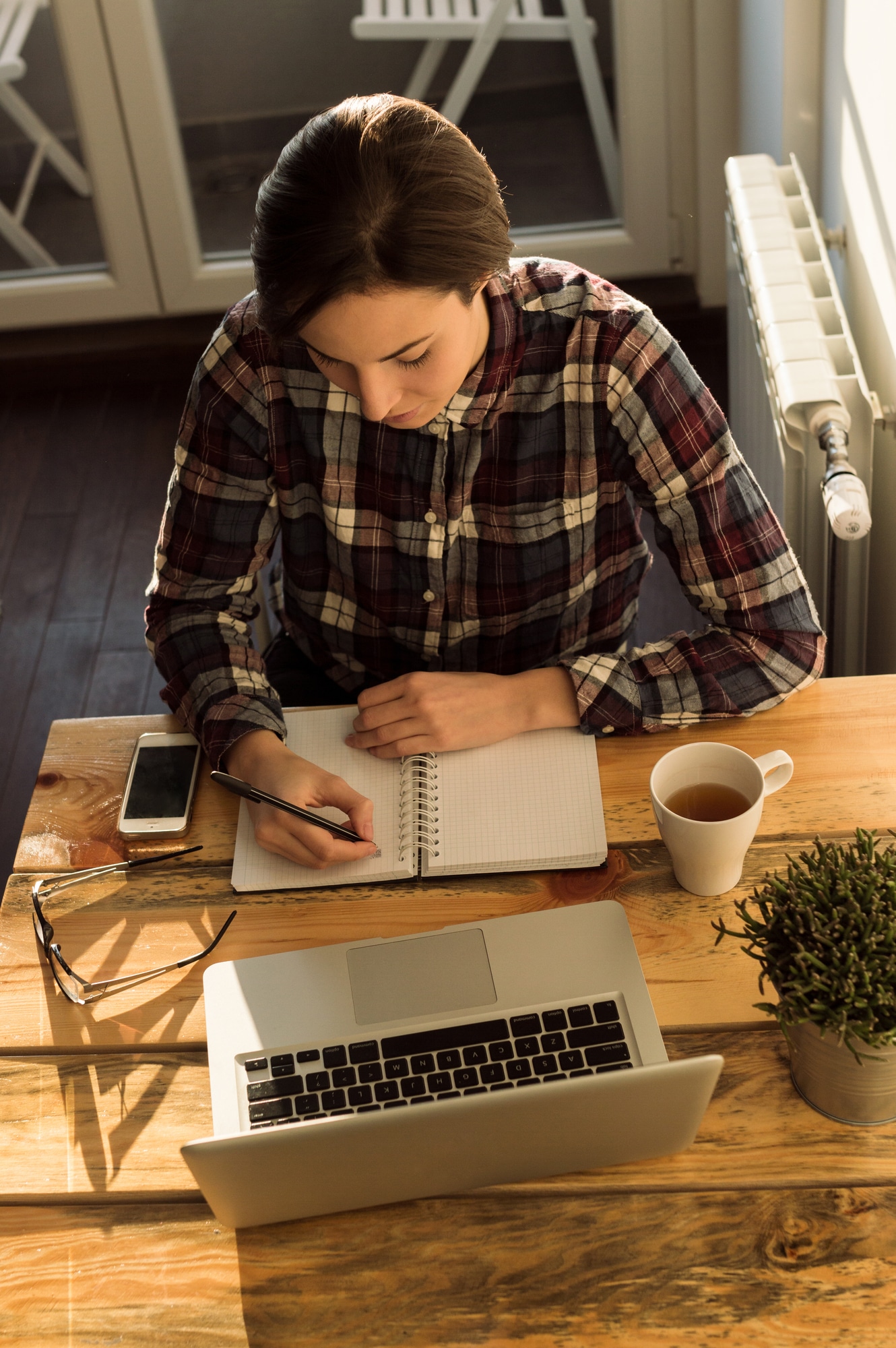 Young female entrepreneur working sitting at a desk working on her laptop computer and notebook in a home office, view from above